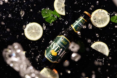 A beer bottle labeled with green and lemon graphics lies on a black surface surrounded by slices of lemon and mint leaves. Water droplets are splashing around, suggesting freshness and dynamism.