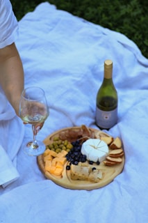 A picnic setup on a white blanket, featuring a wooden platter filled with various cheeses, olives, blueberries, and sliced bread. A hand is holding a glass of white wine, and a bottle of wine is placed nearby. The scene is laid out on a grassy area.