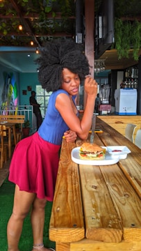 A person leans against a wooden bar table in a colorful café, with a large burger on a white tray in front of them. The café features greenery, hanging lights, and vibrant wall art in the background.