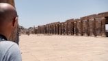 A man is observing an ancient archaeological site with rows of stone columns and a wide paved walkway. The setting is open with clear blue skies and some rocky formations in the background.