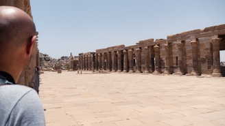 A man is observing an ancient archaeological site with rows of stone columns and a wide paved walkway. The setting is open with clear blue skies and some rocky formations in the background.