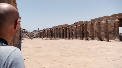 A man is observing an ancient archaeological site with rows of stone columns and a wide paved walkway. The setting is open with clear blue skies and some rocky formations in the background.