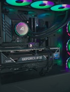 The image depicts the inside of a computer case featuring a high-performance graphics card labeled 'GEFORCE RTX' with RGB lighting. The cooling fans are illuminated with vibrant colors, creating a visually striking look. Various cables and components are neatly arranged, highlighting advanced hardware and a focus on aesthetics.