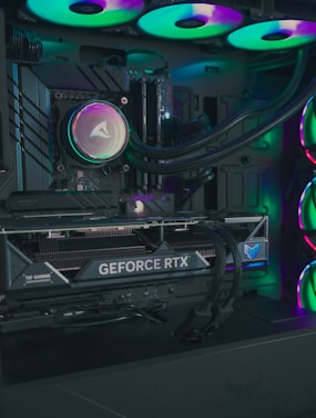 The image depicts the inside of a computer case featuring a high-performance graphics card labeled 'GEFORCE RTX' with RGB lighting. The cooling fans are illuminated with vibrant colors, creating a visually striking look. Various cables and components are neatly arranged, highlighting advanced hardware and a focus on aesthetics.