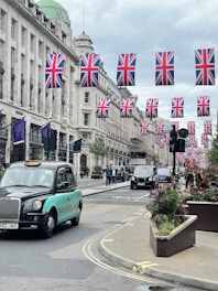 A busy city street lined with tall buildings, decorated with numerous Union Jack flags hanging across the road. Several black taxis and other vehicles are driving down the street. Pedestrians can be seen walking along the sidewalks, and there are large planters with greenery and flowers along the road.