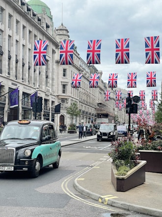 A busy city street lined with tall buildings, decorated with numerous Union Jack flags hanging across the road. Several black taxis and other vehicles are driving down the street. Pedestrians can be seen walking along the sidewalks, and there are large planters with greenery and flowers along the road.