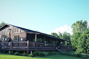 A rustic log cabin with a wooden deck surrounded by lush greenery and trees is seen under a clear blue sky. The cabin has decorative snowflakes on its exterior walls, and various plants can be observed on the deck and around the house.