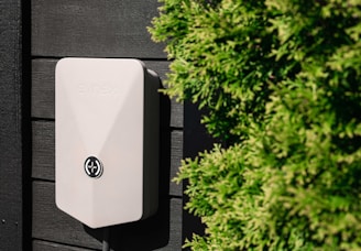 A wall-mounted electrical device with a simple, modern design is affixed to a dark paneled surface. The device has a light gray color and an emblem on its front. On the right side, there are lush green bushes adding a natural element to the scene.