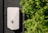 A wall-mounted electrical device with a simple, modern design is affixed to a dark paneled surface. The device has a light gray color and an emblem on its front. On the right side, there are lush green bushes adding a natural element to the scene.