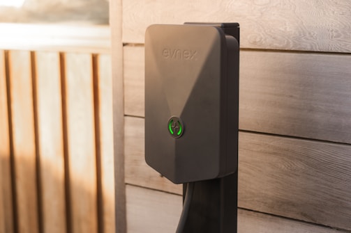 A modern electric vehicle charging station is mounted on a wooden wall. The charger has a sleek design with a matte gray finish and features a green illuminated symbol. The background consists of wood panels with natural grain patterns, suggesting an outdoor installation.