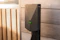 A modern electric vehicle charging station is mounted on a wooden wall. The charger has a sleek design with a matte gray finish and features a green illuminated symbol. The background consists of wood panels with natural grain patterns, suggesting an outdoor installation.