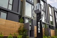 An electric vehicle charging station is positioned in front of a modern building featuring large windows and wooden paneling. The charging unit is mounted on a sleek black stand with a cable wrapped neatly around it. Young plants and greenery are visible at the base of the station.