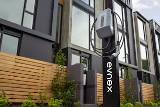 An electric vehicle charging station is positioned in front of a modern building featuring large windows and wooden paneling. The charging unit is mounted on a sleek black stand with a cable wrapped neatly around it. Young plants and greenery are visible at the base of the station.