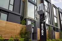 An electric vehicle charging station is positioned in front of a modern building featuring large windows and wooden paneling. The charging unit is mounted on a sleek black stand with a cable wrapped neatly around it. Young plants and greenery are visible at the base of the station.