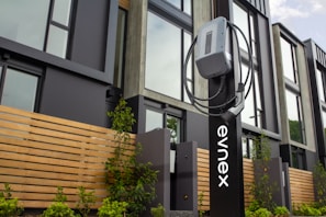 An electric vehicle charging station is positioned in front of a modern building featuring large windows and wooden paneling. The charging unit is mounted on a sleek black stand with a cable wrapped neatly around it. Young plants and greenery are visible at the base of the station.