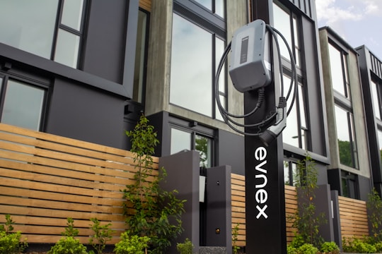An electric vehicle charging station is positioned in front of a modern building featuring large windows and wooden paneling. The charging unit is mounted on a sleek black stand with a cable wrapped neatly around it. Young plants and greenery are visible at the base of the station.