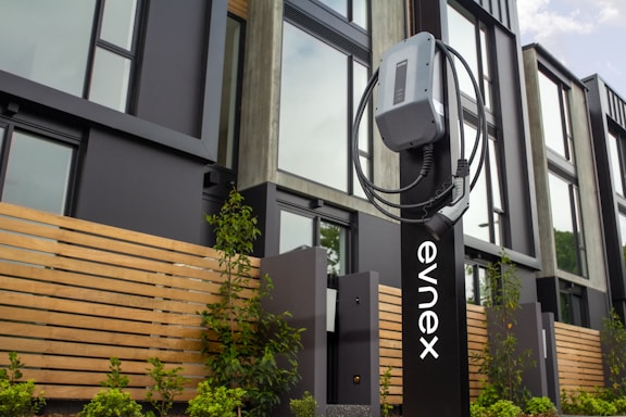 An electric vehicle charging station is positioned in front of a modern building featuring large windows and wooden paneling. The charging unit is mounted on a sleek black stand with a cable wrapped neatly around it. Young plants and greenery are visible at the base of the station.