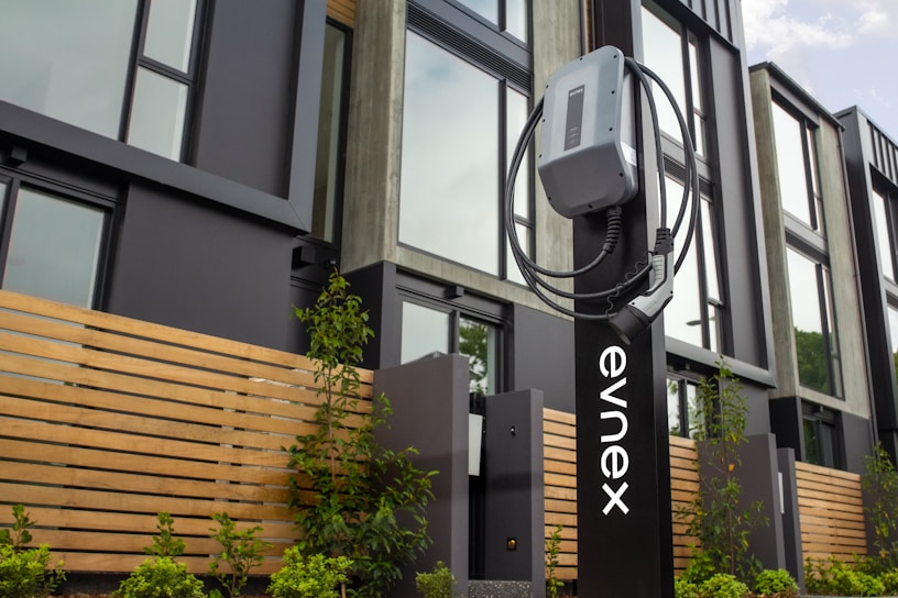 An electric vehicle charging station is positioned in front of a modern building featuring large windows and wooden paneling. The charging unit is mounted on a sleek black stand with a cable wrapped neatly around it. Young plants and greenery are visible at the base of the station.