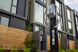 An electric vehicle charging station is positioned in front of a modern building featuring large windows and wooden paneling. The charging unit is mounted on a sleek black stand with a cable wrapped neatly around it. Young plants and greenery are visible at the base of the station.