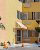 A yellow building facade features several windows with air conditioning units mounted below them. An entrance with a glass door is adorned with a striped awning. The entrance area is bordered by traffic cones and a decorative plant sculpture resembling a hedgehog on the ground nearby. A small manicured tree is visible to the right, and a palm tree can be seen on the left.