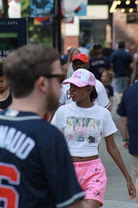 A busy urban street scene with people walking. A woman in the foreground wears pink shorts and a white cropped t-shirt featuring a graphic of three women. She also has a pink cap. Other individuals wearing casual clothing are visible in the background. The setting appears to be a pedestrian area with shops or buildings on either side.