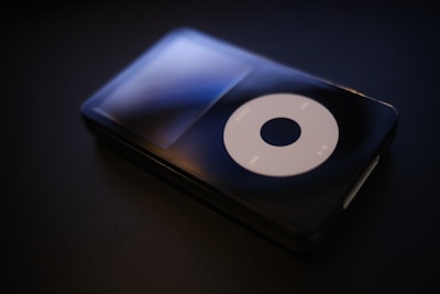 A sleek and glossy black portable music player with a large circular control pad displaying play, pause, and menu buttons, set against a dark background.