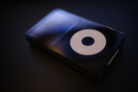 A sleek and glossy black portable music player with a large circular control pad displaying play, pause, and menu buttons, set against a dark background.