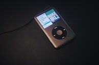 A classic portable music player with a black scroll wheel and a small screen displaying a menu and album cover. It is connected with a cable and rests on a dark surface.