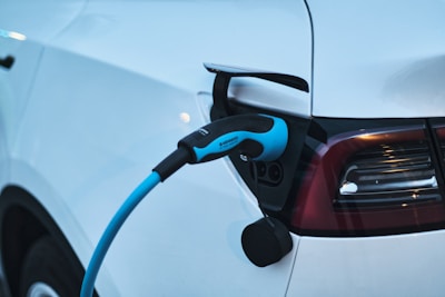 A close-up of an electric vehicle being charged. The charging cable is plugged into the car's port, and the vehicle's taillights and sleek white body are visible.