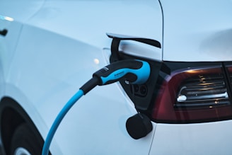 A close-up of an electric vehicle being charged. The charging cable is plugged into the car's port, and the vehicle's taillights and sleek white body are visible.