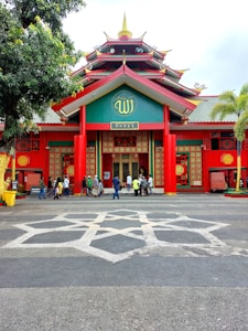 A vibrant building with a pagoda-style roof, featuring red pillars and walls decorated with intricate patterns. A symbol is prominently displayed above the entrance. Several people are gathered near the entrance, and there are palm trees on one side.