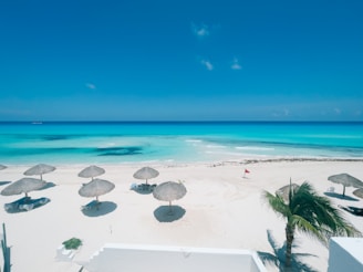 A pristine beach scene showcasing white sandy shores with several straw parasols casting shadows. Crystal-clear turquoise waters stretch out to the horizon under a bright blue sky. A few white lounge chairs are scattered near the parasols, and a red flag waves in the gentle breeze. A lone palm tree is visible on the right side, adding to the tropical ambiance.