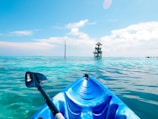 A vibrant blue kayak is floating in clear, turquoise water under a bright, sunny sky. In the distance, there is a small wooden structure with a thatched roof, appearing to be some kind of lookout or watchtower. The kayak paddle is partially visible on the left side of the kayak.