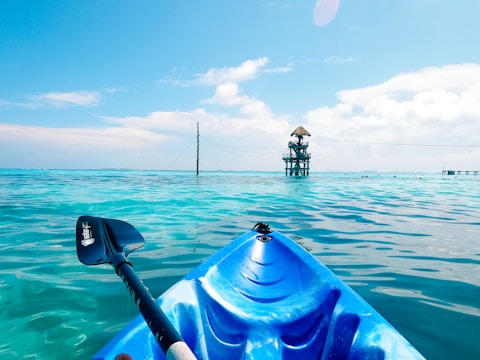 A vibrant blue kayak is floating in clear, turquoise water under a bright, sunny sky. In the distance, there is a small wooden structure with a thatched roof, appearing to be some kind of lookout or watchtower. The kayak paddle is partially visible on the left side of the kayak.