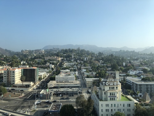 Aerial view of an urban landscape featuring a mix of residential and commercial buildings. The Hollywood Tower building is prominently visible with its sign clearly displayed. Hills and trees are visible in the background under a clear sky.