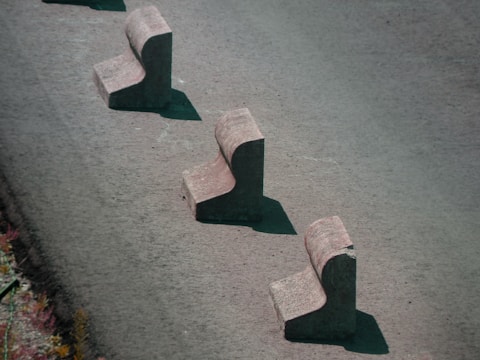 Three concrete barriers are aligned in a staggered pattern on a paved surface. Each barrier has a chunky, L-shaped design, casting shadows on the ground.