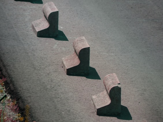 Three concrete barriers are aligned in a staggered pattern on a paved surface. Each barrier has a chunky, L-shaped design, casting shadows on the ground.