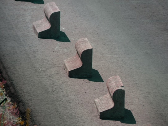Three concrete barriers are aligned in a staggered pattern on a paved surface. Each barrier has a chunky, L-shaped design, casting shadows on the ground.