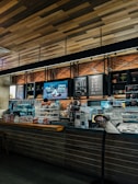 A cozy coffee shop interior with a modern design. The counter is adorned with a variety of pastries in a glass display case. The wall behind the counter has a menu board and decorative shelving with plants. Warm lighting accentuates the brick wall and wooden ceiling.