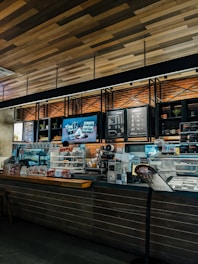 A cozy coffee shop interior with a modern design. The counter is adorned with a variety of pastries in a glass display case. The wall behind the counter has a menu board and decorative shelving with plants. Warm lighting accentuates the brick wall and wooden ceiling.