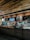 A cozy coffee shop interior with a modern design. The counter is adorned with a variety of pastries in a glass display case. The wall behind the counter has a menu board and decorative shelving with plants. Warm lighting accentuates the brick wall and wooden ceiling.