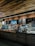 A cozy coffee shop interior with a modern design. The counter is adorned with a variety of pastries in a glass display case. The wall behind the counter has a menu board and decorative shelving with plants. Warm lighting accentuates the brick wall and wooden ceiling.