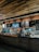 A cozy coffee shop interior with a modern design. The counter is adorned with a variety of pastries in a glass display case. The wall behind the counter has a menu board and decorative shelving with plants. Warm lighting accentuates the brick wall and wooden ceiling.