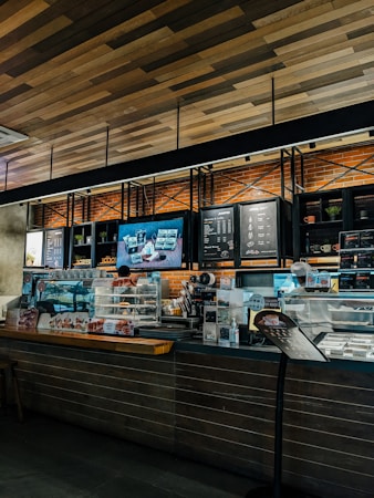 A cozy coffee shop interior with a modern design. The counter is adorned with a variety of pastries in a glass display case. The wall behind the counter has a menu board and decorative shelving with plants. Warm lighting accentuates the brick wall and wooden ceiling.