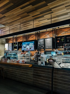 A cozy coffee shop interior with a modern design. The counter is adorned with a variety of pastries in a glass display case. The wall behind the counter has a menu board and decorative shelving with plants. Warm lighting accentuates the brick wall and wooden ceiling.