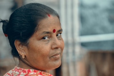 A woman with a contemplative expression gazes over her shoulder. She wears traditional earrings and a vibrant red bindi on her forehead, with her hair neatly tied back. Her attire features shades of red and white, and the background is blurred, suggesting an outdoor or natural setting.