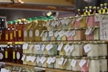 Glass jars filled with colorful substances are neatly arranged on wooden shelves along with bottles of honey. Labels with tags are attached to the jars and bottles, suggesting they are homemade or artisanal products. The jars contain pastel-colored substances, possibly candles or preserved items. The setting appears to be a market or a store.