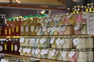 Glass jars filled with colorful substances are neatly arranged on wooden shelves along with bottles of honey. Labels with tags are attached to the jars and bottles, suggesting they are homemade or artisanal products. The jars contain pastel-colored substances, possibly candles or preserved items. The setting appears to be a market or a store.
