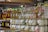 Glass jars filled with colorful substances are neatly arranged on wooden shelves along with bottles of honey. Labels with tags are attached to the jars and bottles, suggesting they are homemade or artisanal products. The jars contain pastel-colored substances, possibly candles or preserved items. The setting appears to be a market or a store.
