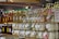 Glass jars filled with colorful substances are neatly arranged on wooden shelves along with bottles of honey. Labels with tags are attached to the jars and bottles, suggesting they are homemade or artisanal products. The jars contain pastel-colored substances, possibly candles or preserved items. The setting appears to be a market or a store.